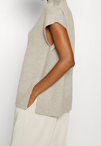Gilet en tricot sans manches couleur beige avec un col haut, bords côtelés, ouvertures sur les côtés et une coupe relaxée, associé à un pantalon de couleur claire.