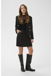 Černý tweedový blejzr se zlatými knoflíky, doplněný o odpovídající černou tweedovou sukni. Modelka má na sobě černé kožené kozačky ke kolenům.