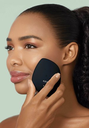 Woman with smooth skin holds a black facial device to her cheek, likely using it for skincare or massage.