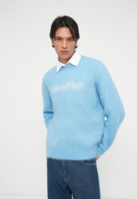 Világoskék kötött pulóver, puha textúrával, kerek nyakkivágással és a Calvin Klein logóval világosabb árnyalatban. Sötét farmernadrággal párosítva.