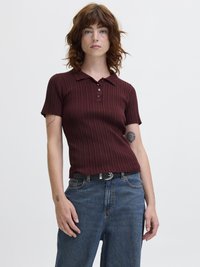 Ung kvinne med bølgete rødt hår har på seg en rillede, kortermet burgunder polo-skjorte, blå jeans, svart belte og viser frem tatoveringer på begge armer.