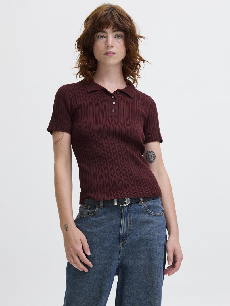 Ung kvinne med bølgete rødt hår har på seg en rillede, kortermet burgunder polo-skjorte, blå jeans, svart belte og viser frem tatoveringer på begge armer.