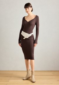 Vestido largo de manga larga color marrón con cuello en V, ceñido en la cintura con un cinturón ancho blanco que tiene agujeros y una hebilla. Combinado con botines beige.