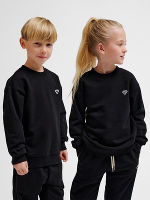 To børn iført sorte matchende sweatshirts og bukser, står side om side mod en ensfarvet baggrund.