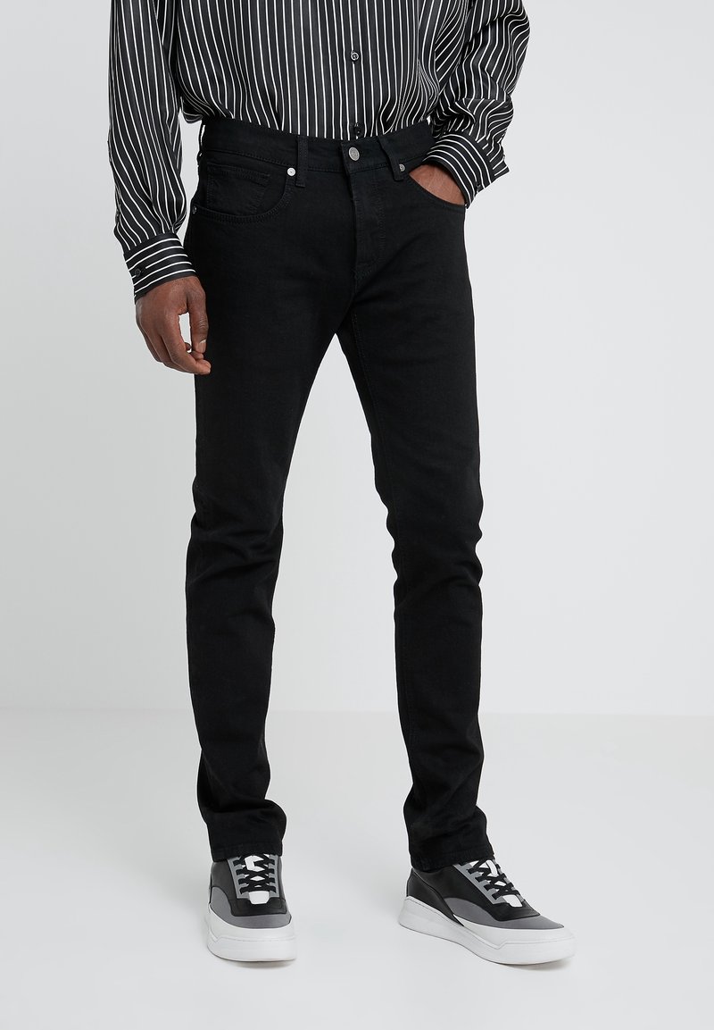 Hombre vistiendo jeans ajustados negros, zapatillas casuales negro y blanco, y una camisa de botones de rayas verticales en blanco y negro.