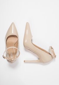 Beiga lacklädershögklackade skor med spetsiga tår och ankelsnören, visade från ovan och sidan mot en vit bakgrund.