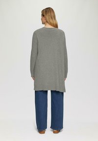 Grijze gebreide lange cardigan met een ontspannen fit, verlaagde schouders en zijsplitsen. Gepaarde met blauwe jeans met een wijd been en bruine schoenen.