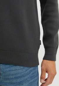 Mørkegrå strikket sweater med ribbede manchetter og kant. Har en tekstureret overflade og et lille logomærke nær bunden. Båret med blå jeans.