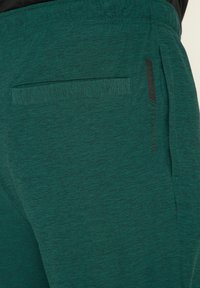 Groene, gestructureerde sportbroek met een achterzak en een zwart logo-accent op de zijkant, ontworpen voor comfort en mobiliteit.