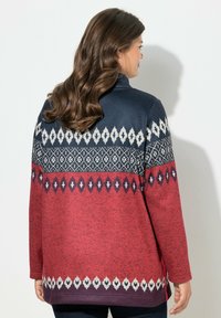 Maglione lavorato a maglia con una parte superiore di colore blu scuro decorata da motivi geometrici bianchi e rossi, che sfuma in una sezione inferiore di colore rosso testurizzato.