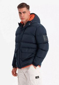 Chaqueta acolchada azul marino con forro de capucha naranja, cierre de cremallera, dos bolsillos frontales y un parche texturizado en la manga izquierda. Diseño cálido y acolchado.