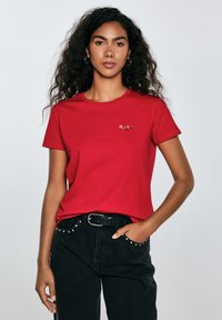Raudona medvilninė t-shirt su apvaliu kaklu ir trumpomis rankovėmis, turinti mažą išsiuvinėtą dizainą kairėje krūtinės pusėje. Dėvima su juodomis džinsomis.
