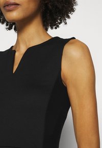 Musta hihaton toppi, jossa on V-mallinen kaula-aukko, rakenteelliset olkapäät ja sileä rakenne, korostaen istuvaa muotoilua.
