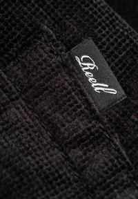 Schwarzer Stoff mit strukturiertem Waffelmuster und genähtem Rand. Ein kleines Etikett mit weißem Text ist sichtbar und lautet "Reell."