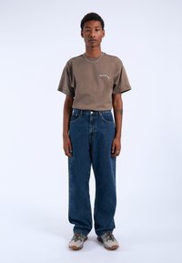 T-shirt marrone a maniche corte con un piccolo logo, abbinato a jeans di denim blu larghi e sneakers grigie a fantasia. Sfondo semplice.