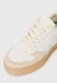 Hvit skinnsneaker med beige semsket detaljer, rund tå, perforerte detaljer og kremfargede lisser, med en teksturert gummisul.