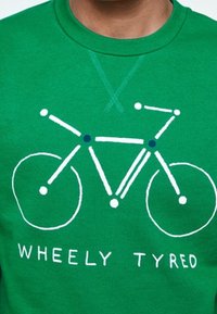 Zelená mikina s grafickým motivem bicyklu obrysovaného bílou barvou, s modrými kruhovými akcenty a textem "WHEELY TYRED" pod ním.