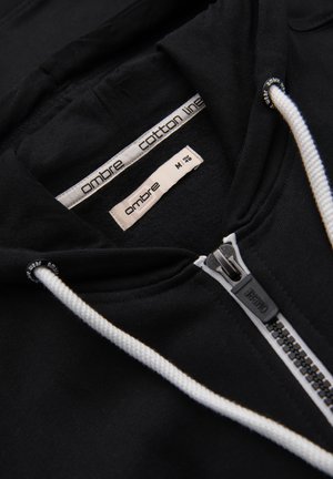 Schwarzer Kapuzenpullover mit Reißverschluss, weißen Kordeln, einem Reißverschlussanhänger mit Markenaufdruck und Etiketten im Kragen mit der Aufschrift "ombre cotton line" und Größe "M".