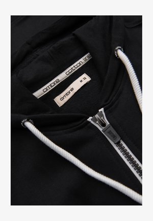 Schwarzer Kapuzenpullover mit Reißverschluss, weißen Kordeln, einem Reißverschlussanhänger mit Markenaufdruck und Etiketten im Kragen mit der Aufschrift "ombre cotton line" und Größe "M".