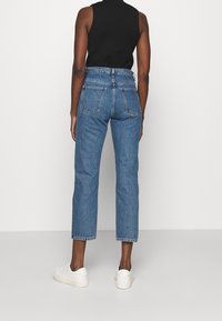 Jeans de mezclilla azul con una cintura alta, pierna recta, cinco bolsillos y un acabado desgastado. Llevados con una camiseta negra sin mangas y zapatillas blancas.