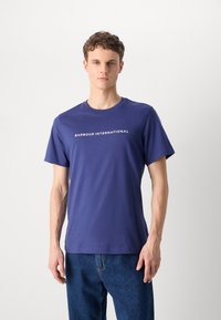 Navy blue cotton t-shirt with short sleeves and a crew neck, featuring white text "BARBOUR INTERNATIONAL" on the front. Model wears blue jeans.