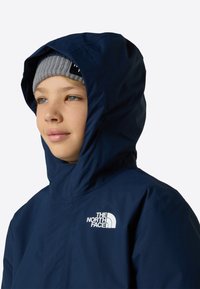 Jachetă bleumarin cu glugă și finisaj neted, cu un logo pe piept. Poartă o caciulă tricotată gri deschis.