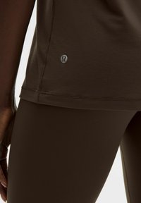 Braunes Sportoberteil mit kurzen Ärmeln und kleinem Logo am Saum. Der Stoff wirkt glatt und wird mit passenden braunen Leggings kombiniert.