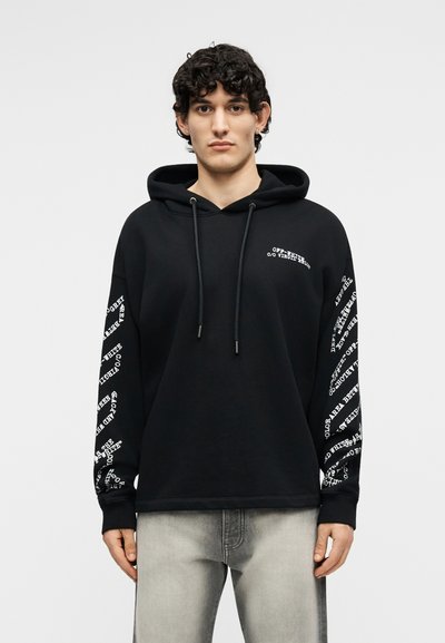 Jeune homme aux cheveux bouclés portant un sweat à capuche noir avec du texte blanc sur les manches et la poitrine, associé à un jean gris clair, debout.