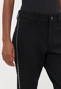 Zwarte broek van rekbare stof, voorzien van hartvormige studs langs de zijkant, met een platte tailleband en een enkele knoopsluiting.