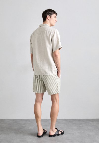 Camicia di lino beige chiaro a maniche corte abbinata a pantaloni corti a righe verdi e bianche, indossata con sandali neri. La camicia presenta un colletto classico.