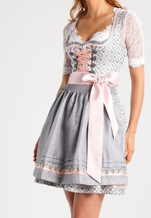Kobieta w tradycyjnej bawarskiej sukience dirndl z koronkowymi rękawami, różową wstążkową gorsetą i kratkowym fartuszkiem zawiązanym dużą różową kokardą.