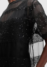 Robe noire en tissu transparent ornée de sequins décoratifs disposés en rayures horizontales. La texture varie entre des zones lisses et des zones embellies.