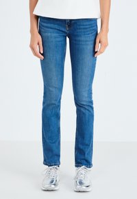 Kék farmernadrág, slim fit fazonnal és közepes mosással, varrási részletekkel; ezüst sportcipővel és fehér felsővel viselve.