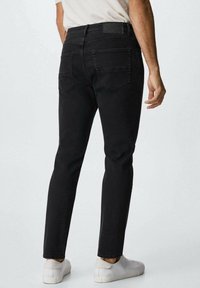 Hombre vestido con jeans negros ajustados, zapatillas blancas y una camisa blanca, de pie contra un fondo claro liso, mostrado desde atrás.