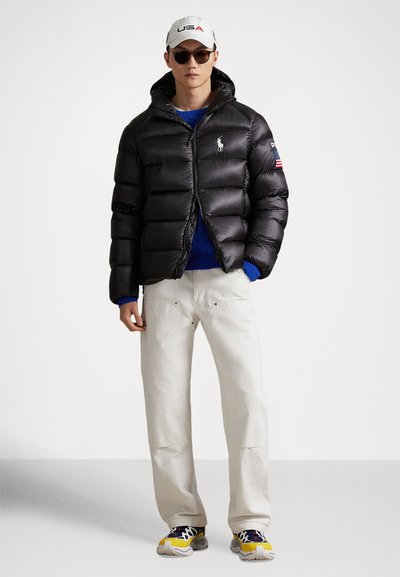 Fekete steppelt dzseki levehető kapucnival, vízszintes varratokkal és logóval. Kék pulóver fölött viselve, krémszínű nadrággal és színes sportcipőkkel párosítva.