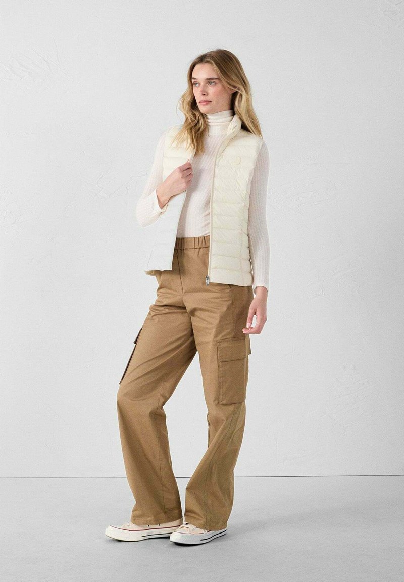 Gilet smanicato avorio sopra un dolcevita aderente, abbinato a pantaloni cargo marroni larghi con tasche laterali e sneakers bianche.