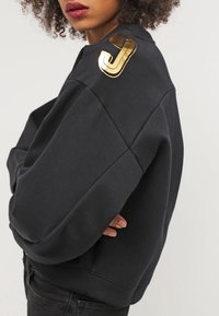 Sweat-shirt noir oversize en tissu texturé, avec un accent doré brillant en forme de lettre "J" sur l'épaule.