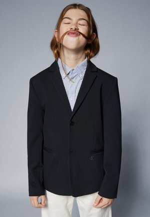 Blazer negro con un solo botón, con dos bolsillos frontales y un logo sutil. Combinado con una camisa a rayas azul y blanca.