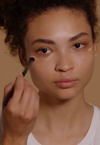 Application de maquillage à l'aide d'un pinceau, en se concentrant sur le contour des yeux. Le sujet a la peau de ton moyen et des cheveux bouclés, portant un haut blanc.