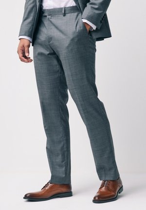 Man wearing tailored grey suit pants with hand in pocket and polished brown leather dress shoes on white background.
