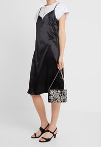 Černé saténové šaty na ramínka nošené přes bílé tričko, doplněné černými sandály na vysokém podpatku a černou zdobenou kabelkou.