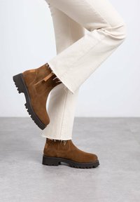 Botines de ante marrón con suela de goma negra gruesa y cremallera lateral, combinados con unos pantalones vaqueros blancos rotos por el bajo.