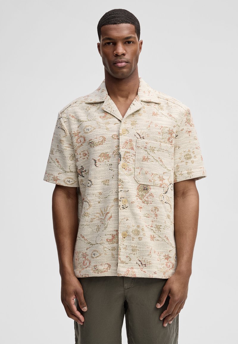 Uomo che indossa una camicia beige a maniche corte con bottoni, caratterizzata da un sottile motivo astratto multicolore e una tasca frontale, in piedi davanti a uno sfondo semplice.