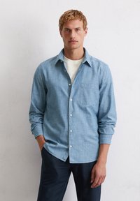 Chemise bleu clair à manches longues en tissu chambray, dotée d'une seule poche poitrine. Portée avec un sous-vêtement blanc uni.