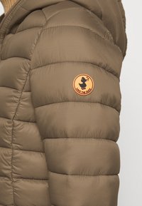 Gros plan sur la manche d'une veste matelassée marron avec un patch rond orange montrant une silhouette de canard et le texte "SAVE THE DUCK."