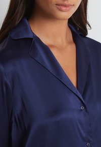 Chemise satinée marine avec col en V, fermeture à boutons et texture lisse. Le tissu a une finition brillante qui accentue son élégance.