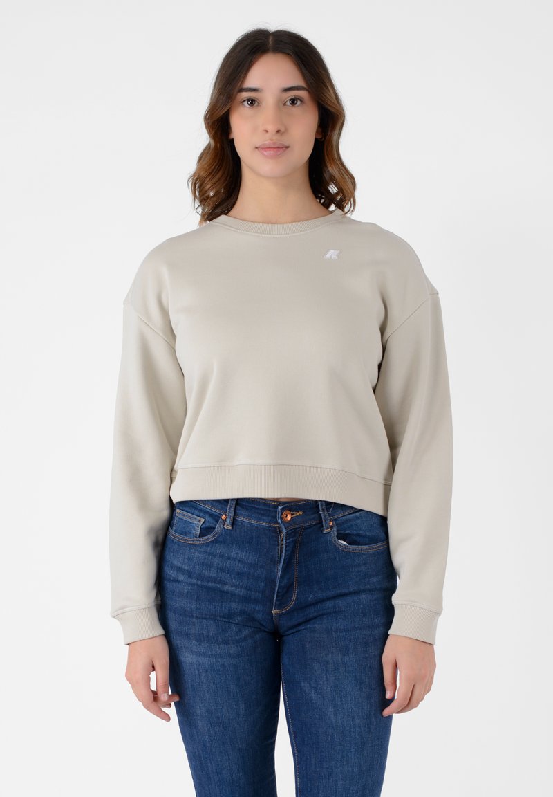 Felpa beige tagliata con scollatura rotonda, polsini a coste e un piccolo logo sul petto, abbinata a jeans blu.