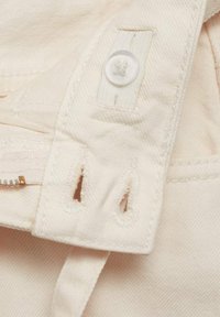 Tissu beige avec une texture lisse, doté d'un bouton blanc, d'une fermeture éclair avec des accessoires en or, et de petites découpes pour l'attache.