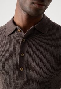 Camisa polo de punto acanalado marrón con cuello tipo polo, tapeta de tres botones y textura suave. Presenta botones de color natural.