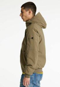 Giovane uomo in giacca con cappuccio color beige, con tasca con bottone sulla manica, camicia gialla e jeans blu, mostrato di profilo su uno sfondo bianco.